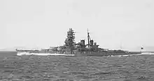 Side view of a dark gray warship at speed. She has two funnels, one mast and a massive forward superstructure.