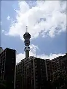 A concrete tower, the bottom obscured by other buildings, viewed from below. A large football shaped structure has been fitted to the tower just below the habitable section.