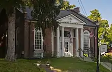 A small brick building in a neoclassical style, with two columns beside the main entrance and a gabled roof, seen from its front right, in the shade of a tall spruce tree. A sign next to the steps up from the street says "Hillsdale Town Hall".