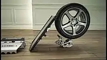 a flat board strikes the tread of a car tyre at a 45-degree angle. The tyre is balanced on top of a smaller piece of machinery on a wooden floor.