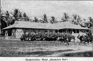 A small single story building, with grass roof and veranda, next to bushes