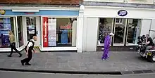 A man in a purple jumpsuit is at the right of the image walking down a street. On the left is a man dressed as a police officer following him. At the far right is a man seated on the back of a golf cart filming them. Storefronts can be seen in the background.