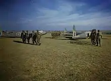 Three groups of six men wearing helmets and backpacks walk across a grass field towards waiting aircraft