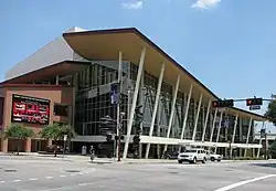 Concrete auditorium fronted by a large glass atrium over which is a slightly curved roof supported by narrow, angled poles.