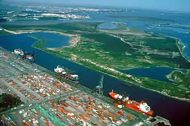 A large paved area appears in the foreground with many rows of cargo and storage facilities. It is adjacent to a ship channel with three docked freighters visible. In the background one sees the ship channel is part of a larger network of small bays at the mouth of Buffalo Bayou.