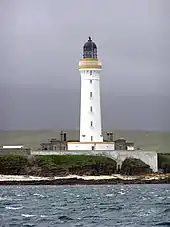  A tall white lighthouse with a brown stripe around the parapet and dark coloured lantern sits on a rocky shore. A white wall obscures the lower floor of grey stone buildings gathered around its base.