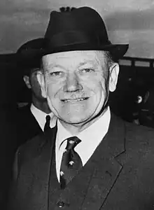 A middle-aged man wearing a dark suit and tie, and a hat. He has a toothbrush moustache.