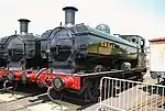 Two pannier tank locomotives are seen from the front and to the right and are standing next to each other in front of a turntable. Both locomotives have black chimneys and fronts. The cabs, pannier tanks, clackbox covers and boiler domes are all green. The buffer beams are red and include the locomotive number in yellow with black edging, 7752 on the left locomotive and 7760 on the right. On the right locomotive, one round cab window is visible and is edged in brass.