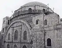 A large domed roof sits upon a bricked parapet, which includes arched windows, three of which are visible. The base of the parapet is surrounded by a narrow veranda, which is fenced with decorative metal grating. Below this, a set of three large arched windows, on two levels, are situated under a large stone arch, itself flanked by two supporting towers. The left tower consisting of an upper section is slightly taller that its counterpart on the right.