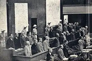 Three rows of benches with a dozen or so men standing behind each. Behind them stand five men in uniform.