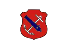 An insiginia in the form of a red shield. On the shield are a white anchor crossed by a blue cannon barrel.