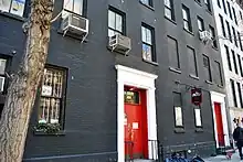 A black-brick multi-storied building with red doors and a tree outside it.