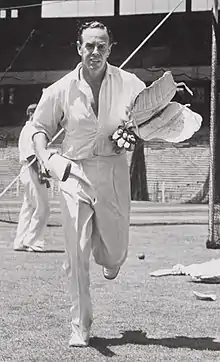 Johnson, bare-headed and wearing cricket whites, running towards the camera carrying cricket pads and gloves in his left hand and a cricket bat in his right hand