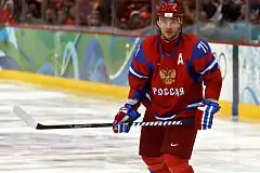 An ice hockey player is standing while slightly turned to his left.  He has short dark hair and is not wearing a helmet.  He is wearing a blue uniform with a large orange bird with an ice hockey stick on his chest.