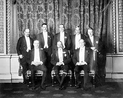 A monochrome photograph of men wearing white tie with breeches, three of them (Baldwin for the United Kingdom, George V, and King for Canada, from viewer's left to right) seated in the front row and five (Monroe for Newfoundland, Coates for New Zealand, Bruce for Australia, Hertzog for South Africa, and Cosgrave for Ireland, ditto) standing behind them.