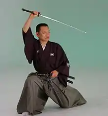 A man in baggy robes and split pants kneels on one knee and holds a Japanese sword above his head.