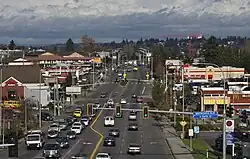 An aerial view of a suburban street with several traffic lanes and traffic lights amid strip malls and gas stations.