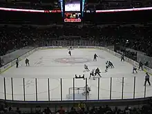 An ice hockey rink on which two teams are playing seen from some distance up and behind the goal. High transparent shielding mounted on the wall surrounds the rink.