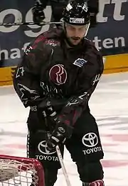 An ice hockey player standing directly in front of the camera. He is wearing a black helmet with a visor and a black and red uniform.