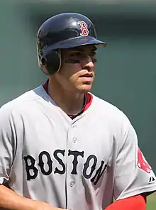 A young man in a grey baseball jersey wearing a batting helmet with eyeblack under his eyes.