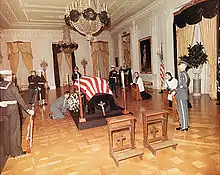 casket draped with a flag in a large room.  Several soldiers stand around it, and two clerics kneel on the kneelers at the side.  Two empty kneelers are in the foreground