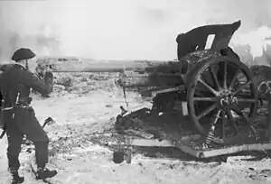 Black-and-white photograph of Churchill in uniform looking down the barrel of a large artillery gun with A stone barricade visible in the background