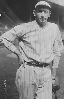 A man in a white baseball uniform with dark pinstripes and a cap wears a baseball glove on his left hand as he leans on a baseball bat with his other hand on his hip.