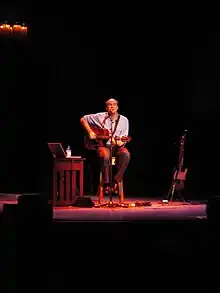 A man behind a microphone holding an acoustic guitar, sitting on a stool in the center of a stage, with a light shining down from above. To his sides are an electric guitar on a stand, a side table with a laptop, a bottle, and some additional equipment.