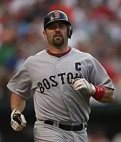 baseball player standing in a grey uniform with a letter C on his left chest and the letters "BOSTON" visible on his jersey. He is jogging with a navy batting helmet on.