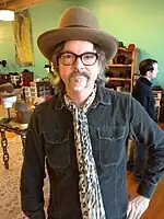 A man with a goatee stands in a room with his left hand on his hip. He is wearing a brown fedora, dark-brown glasses, a pocketed dark-teal shirt, and a leopard-print scarf.