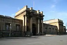 A stone building on an asphalt street. A wrought iron fence separates the building and street. Four columns flank the entrance, a high archway over which is chiseled "OXFORD UNIVERSITY PRESS". Architectural wings extend from either side of the main section of the building. A bright blue sky is visible in the background.