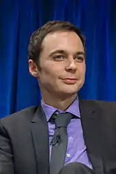 A brunette man wearing a purple dress shirt, black tie, and black jacket stares off into the distance with a slight smirk