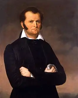 Three-quarter portrait of a young clean-shaven man with long sideburns and a widow's peak hairline. His arms are crossed.