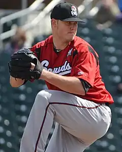 A man wearing a black baseball cap and a red jersey with "Sounds" written across the chest in white script and gray baseball pants shown in the midst of his windup