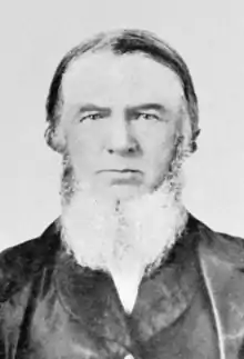 Black and white photograph of John Baker White, who has a beard and is wearing a suit.