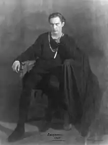 Barrymore, cleanshaven, in an all-black costume as a brooding Hamlet, sitting on a chair, looking slightly to his right of the camera