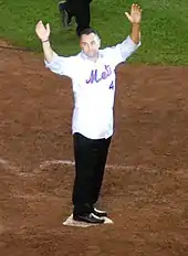 A man in a white baseball jersey and blue jeans stands on home plate with his arms upraised.