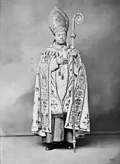 A bespectacled man holding a crosier in his left hand and wearing a mitre and liturgical vestments faces forward