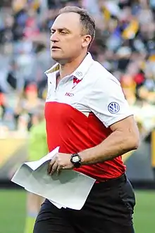 A dark-haired man in a red-and-white polo carrying papers