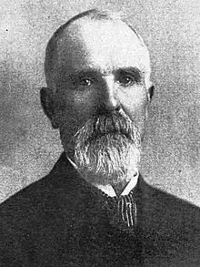 Black-and-white photograph of a man with thin hair and a beard in a suit and tie, with a stern expression