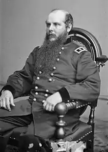 Black and white photo of a seated man in a dark military uniform with two stars on his shoulder tabs. He is balding and has a long beard.