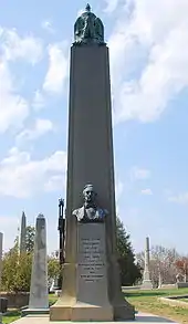 A large obelisk in a graveyard, with a bust of Tyler, and a black cast iron cage partially visible behind it.