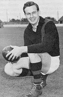 A dark-haired footballer in a long-sleeve black guernsey with a diagonal sash and white shorts squatting while holding a football