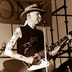 Black & white photo of Johnny Winter from the waist up playing a Gibson Firebird guitar on a stage