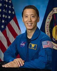 An Asian man in a blue flight suit is sitting in front of US & NASA flags; he is facing the camera and smiling.