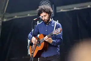 José González wearing a dark blue shirt, standing onstage in front of a microphone, playing an acoustic guitar