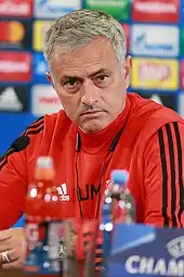 The head and upper torso of a man with black, greying hair. He is wearing a black polo shirt with the blue Chelsea F.C. crest on the left breast and a white logo of the Adidas sponsor on the right breast. Three white stripes are visible on the shoulder.