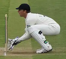 A young man with light brown hair is standing on some grass. He is wearing a burgundy, grey and black cricket uniform, large gloves and black trousers covered by black think pads on his lower legs.