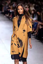 A brunette woman walks down a fashion runway in a tan-colored zipper garment over a black turtleneck dress