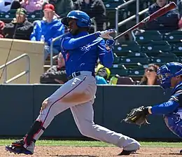 A man wearing blue baseball jersey and gray pants after swinging at a ball stands at home plate